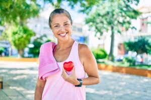 Woman in pink workout shirt and towel holding a toy heart while smiling